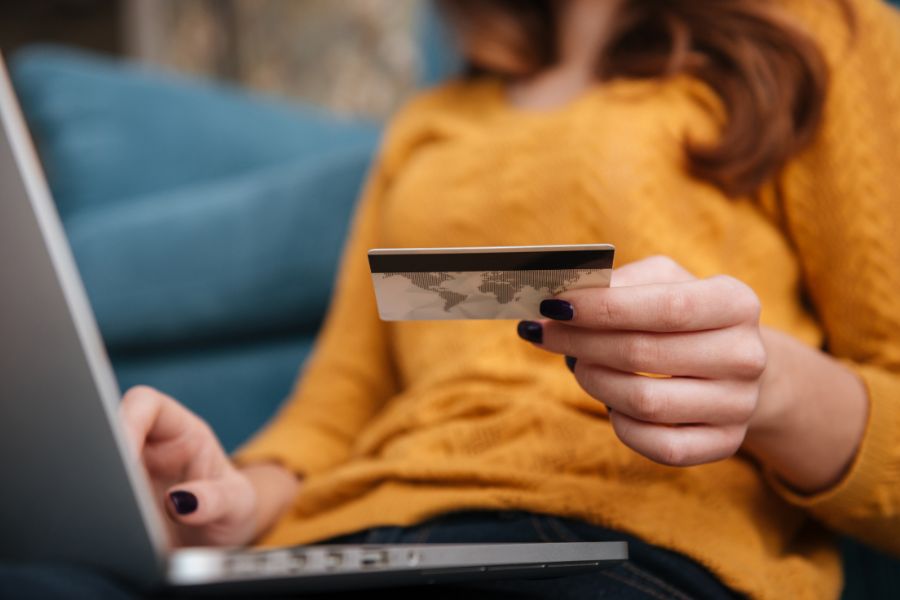 אישה מחזיקה כרטיס אשראי ועל רגלה מחשב נייד.